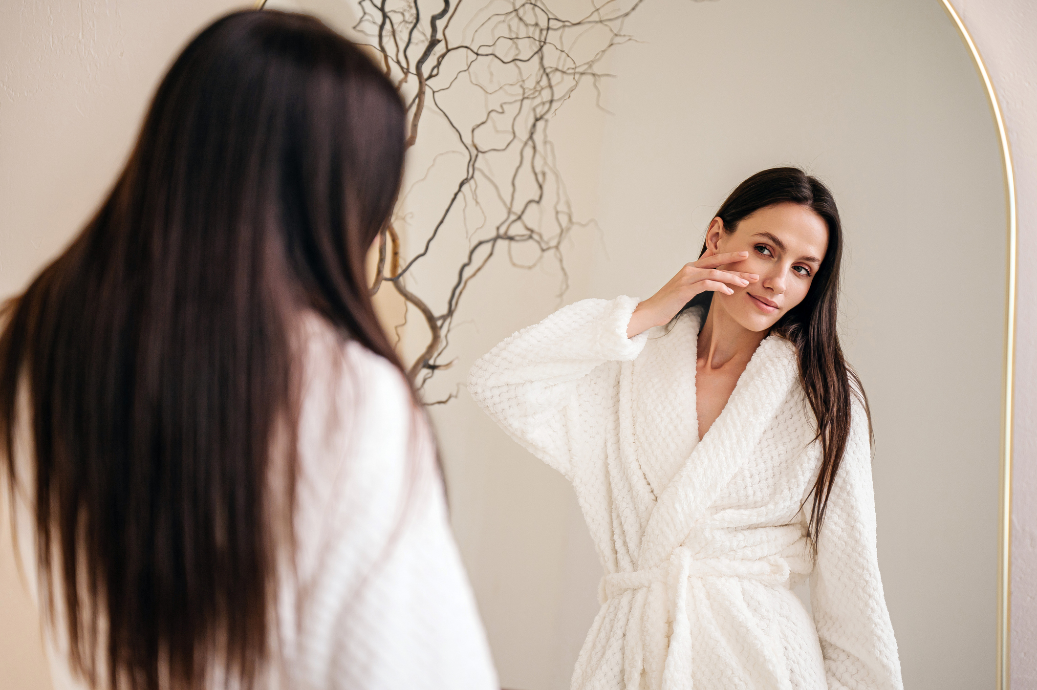 woman in white shower bathrobe touching her face, looking at reflection in mirror, applying face cream. Beauty concepts of skin care routine at home